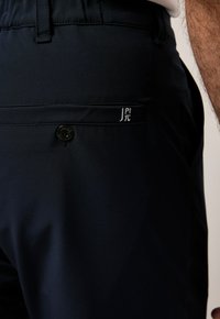Marinblå byxor med en slät yta, med en bakficka med knappstängning och broderad logotyp på sidan.