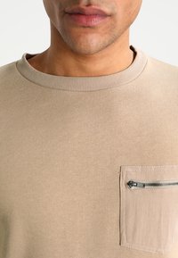 Felpa beige con scollo rotondo, polsini a costine e tasca sul petto con zip. Tessuto morbido e vestibilità casual.