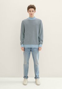 Jasnoniebieski sweter z dzianiny z wysokim kołnierzem, z teksturowanym wzorem, w połączeniu z wyblakłymi niebieskimi dżinsami i białymi trampkami.