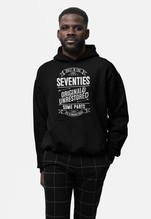 Mann trägt einen schwarzen Hoodie mit weißem Text "In den siebziger Jahren gebaut, original & unrestauriert, einige Teile noch funktionsfähig" und karierte Hose.