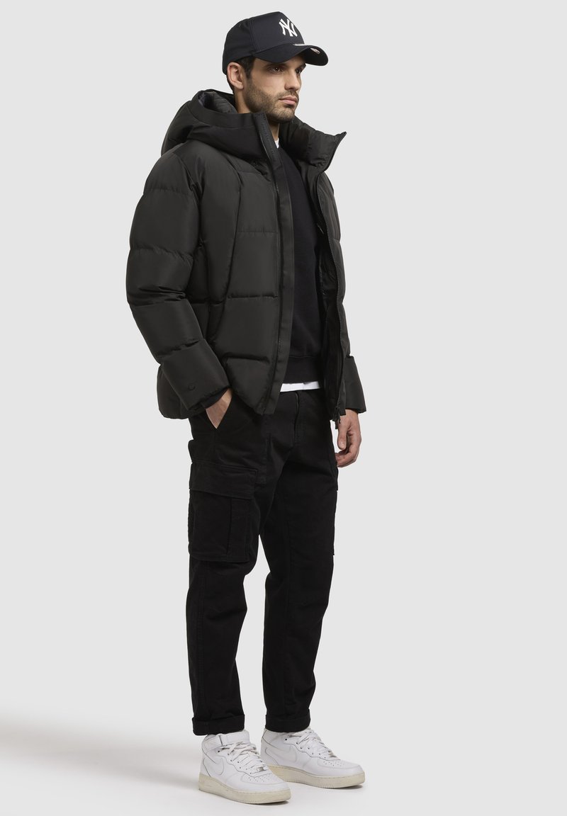 Schwarze Pufferjacke mit Kapuze, Reißverschluss und gestepptem Design. Kombiniert mit schwarzen Cargo-Hosen und weißen High-Top-Sneakern.