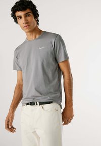 T-shirt grigia in cotone a maniche corte con scollatura rotonda e un piccolo logo bianco sul lato sinistro del petto. Abbinata a pantaloni chiari e una cintura nera.