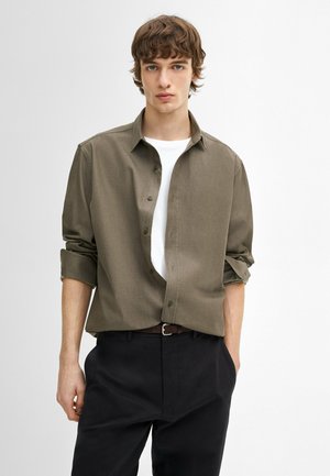 Camicia verde oliva con bottoni, maniche arrotolate, sotto una maglietta bianca a girocollo e pantaloni scuri; include bottoni frontali standard e un colletto.