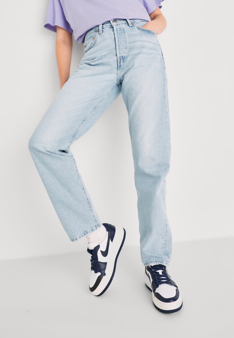 Personne portant un jean taille haute bleu clair, des chaussettes blanches, des baskets noires et blanches, et un haut à manches courtes lavande, debout devant un fond blanc.