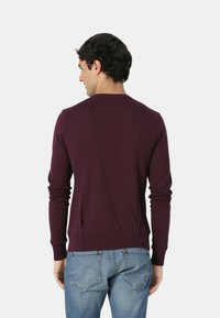 Maglione bordeaux a maniche lunghe con polsini a coste e scollo rotondo, caratterizzato da una texture liscia e un design aderente, indossato con jeans azzurri.