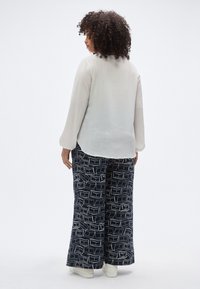 Λευκή μπλούζα με μακριά μανίκια συνδυασμένη με σκούρο μπλε παντελόνι φαρδιάς γραμμής με γεωμετρικό λευκό μοτίβο, και τα δύο κατασκευασμένα από ελαφριά υφάσματα.