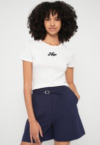 Fehér pamut póló fekete "Hugo" logóval, kerek nyakkivágással, rövid ujjakkal, sötétkék, magas derekú rövidnadrággal párosítva. Simább textúrával.