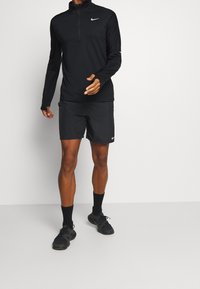 Maglietta nera a maniche lunghe con zip a quarti e pantaloncini abbinati con il logo Nike; vestibilità atletica; tessuto leggero; indossato con calze e scarpe nere.