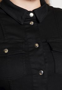 Chemise en denim noir avec col, boutons métalliques et deux poches poitrine. Le tissu a une texture lisse avec des détails de couture contrastants.