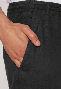 Zwarte linnen shorts met een elastische tailleband en een zijzak, met een close-up van een hand die in de zak rust, waarmee de textuur wordt benadrukt.
