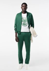 Chándal verde hecho de tela suave con acentos blancos. Presenta una chaqueta con cremallera, una camiseta de cuello redondo con gráfico y zapatillas blancas.