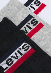 Tre paia di calzini in bianco, grigio e nero, ciascuno con un prominente logo Levi's in rosso e blu sul bordo. Texture lavorata morbida.