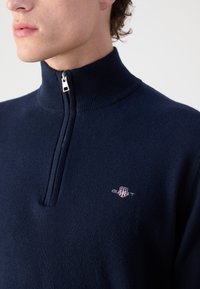 Pulover cu fermoar navy din material tricotat moale, cu guler înalt, accente ribbate și un mic logo brodat pe piept.