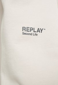 Suéter de algodón en un beige claro con una textura suave, que presenta una impresión negra de "REPLAY" y "Second Life" en la parte frontal.