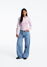 Camicia button-up rosa chiaro con dettaglio arricciato davanti, abbinata a jeans in denim azzurro chiaro a gamba larga. Calzature nere visibili.