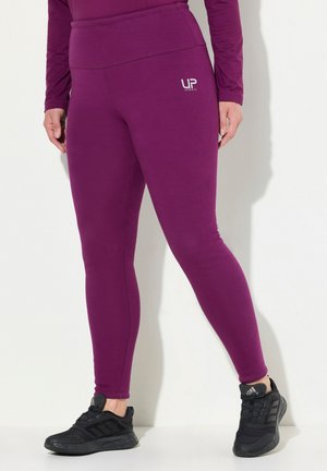 Person in lila UP Sports Leggings und schwarzen Adidas Turnschuhen, steht entspannt mit den Händen an den Seiten gegen eine weiße Wand.