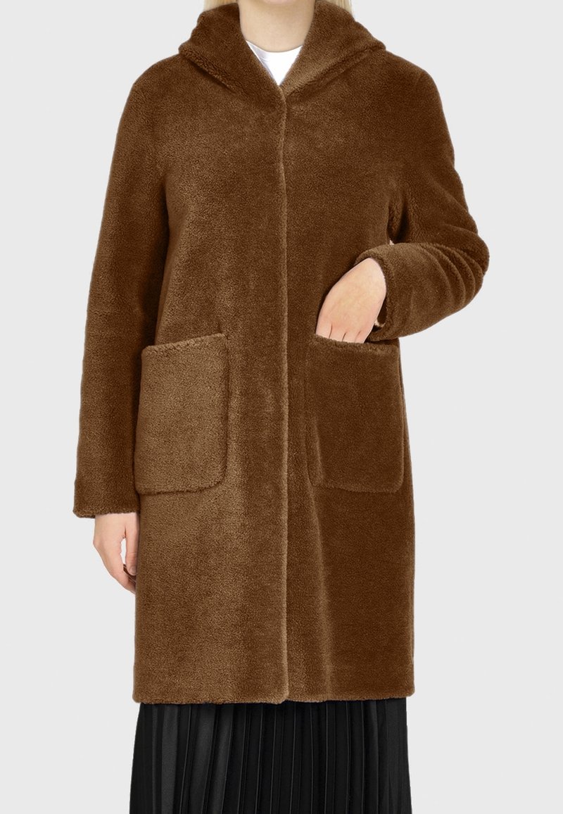 Personne portant un long manteau à capuche en peluche marron avec de grandes poches avant, une main dans une poche, et une jupe plissée noire visible en dessous.