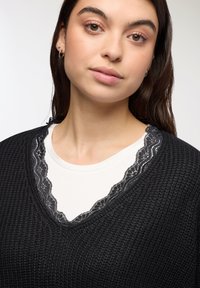 Młoda kobieta z brązowymi oczami i kolczykami, ubrana w czarny sweter dzianinowy nałożony na biały top z czarną koronką.