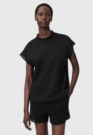 Femme portant un haut noir à manches courtes et un short assorti avec bordure en dentelle, debout devant un fond uni.