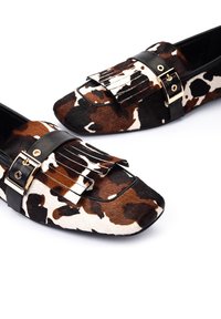 Mocasines de estampado animal con una combinación de material de piel de vaca marrón y blanca. Presentan detalles en cuero negro y herrajes en tono dorado.