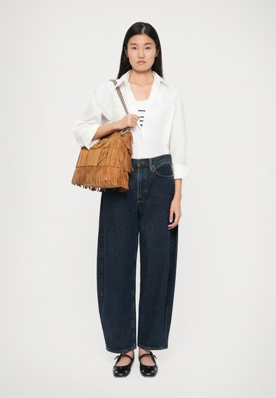 Donna che indossa una camicia bianca, una maglietta a righe, jeans wide-leg scuri e ballerine nere, mentre tiene una grande borsa a tracolla frangiata color beige.