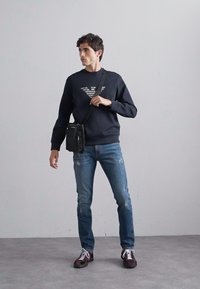 Hanorac bleu marin cu un design grafic, blugi cu spălare medie și detalii deteriorate, și adidași burgundy cu accente albe. Geantă de umăr neagră.