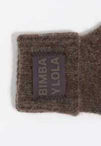 Tela de punto texturizada marrón con un parche de cuero rectangular marrón oscuro que presenta el texto en relieve "BIMBA Y LOLA" en el borde.
