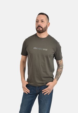 Uomo con capelli corti e barba che indossa una t-shirt verde Armani Exchange e jeans blu, mostra la manica del tatuaggio sul braccio destro, mani nelle tasche.
