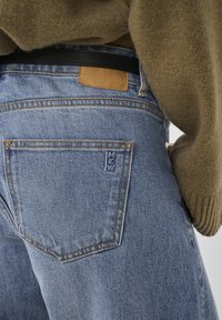 Svetlomodre jeans hlače iz jeansa z rjavim usnjenim ovratnikom v pasu, poudarjenim šivanjem in vidnimi zadnjimi žepi.