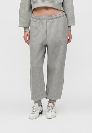 Personne portant un pantalon de survêtement gris à jambes larges, un sweat court et des baskets blanches avec des détails noirs et dorés, debout sur un fond clair.