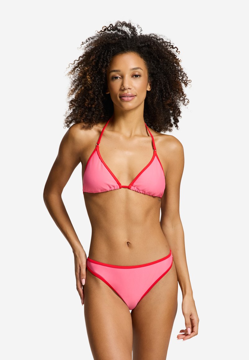 Mujer con cabello rizado que lleva un bikini rosa con ribete rojo, de pie con una mano en la cadera contra un fondo blanco liso.