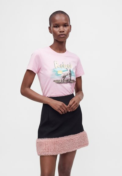 Νεαρή γυναίκα που φοράει ένα ροζ γραφικό t-shirt με το κείμενο "Rebel" και μια μαύρη φούστα με ροζ γούνινη επένδυση, στέκεται μπροστά σε λευκό φόντο.