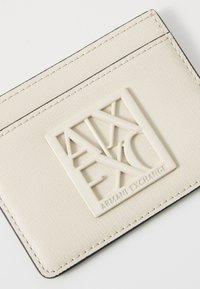 Bijeli teksturirani držač za kartice s istaknutim uzdignutim logom u kvadratnom dizajnu. Šivani rubovi i glatka završna obrada upotpunjuju izgled.