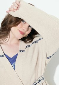 Beige stickad cardigan med blå accentstripes och ett mönstrat midjeband, med v-ringning och öppen framdesign.