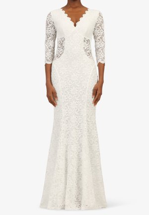 Kobieta ubrana w białą koronkową suknię sięgającą podłogi, z rękawami do połowy przedramienia i dekoltem w kształcie litery V z falbanką.