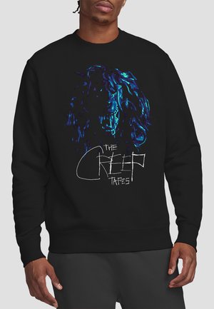 Man wearing black sweatshirt with blue-toned snarling dog graphic and "The Creep Tapes" text on front against gray background.