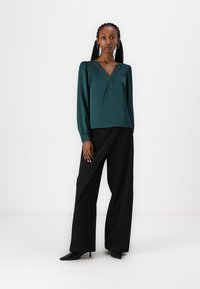 Teal satina blouse dengan garis leher V dan hiasan renda. Dipadukan dengan celana hitam legang berpinggang tinggi. Tekstur halus, lengan panjang.