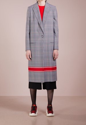 Osoba nosící kabát s károvaným vzorem po kolena s červeným pruhem blízko lemu, černé zkrácené kalhoty, černé punčocháče a červené kostkované tenisky.