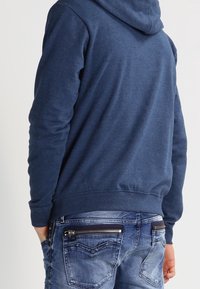 Persona vistiendo una sudadera azul y jeans azul desgastados con bolsillo trasero con cremallera, de pie con las manos en los bolsillos, de espaldas.