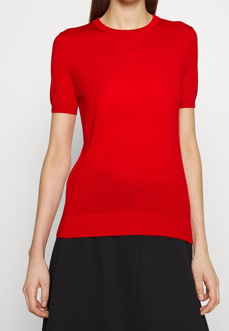 Femme portant un pull rouge à manches courtes ajusté, associé à une jupe noire, vue du cou jusqu'aux hanches sur un fond blanc.