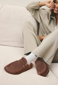 Braune Wildlederschlüpfer mit flauschiger Innenausstattung, mit einer gestickten Kante und einem goldenen Logo. Getragen mit weißen Socken und beiger Freizeitkleidung.