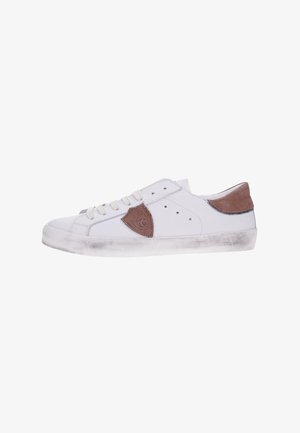 Zapatilla blanca de corte bajo con parches de cuero marrón en el lateral y el talón, diseño con cordones y suela de goma ligeramente gastada.