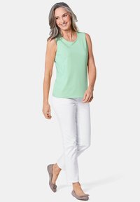 Mintgroene, mouwloze top met een ronde hals, gecombineerd met witte broeken en grijze flats, gepresenteerd op een model in een staande pose.