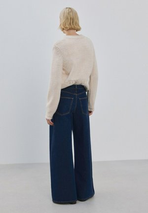 Persona con cabello rubio corto, vistiendo un suéter color crema y jeans anchos azul oscuro, de pie contra un fondo claro y sencillo.