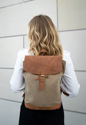 Blonde woman in white shirt wearing tan and brown leather-accented backpack, standing against light gray wall with rectangular panels.