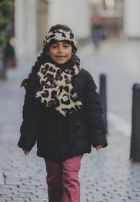 Zwarte pufferjack met een sjaal en hoofdband met luipaardprint, lichtbruine broek, wandelt op een straat met kasseien.