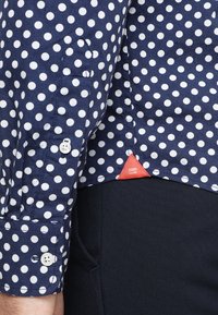 Marineblauwe shirt met witte stippen, met knoopmanchet en een rood driehoekig label boven de zak van de marineblauwe broek.