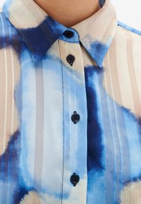 Chemise à col avec des rayures verticales beige et des motifs abstraits bleus en aquarelle, fermée devant par des boutons noirs.
