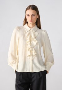 Blusa de color crema, texturizada, con fruncidos en el frente, cuello y mangas largas. Botones en la parte delantera, combinada con pantalones negros.
