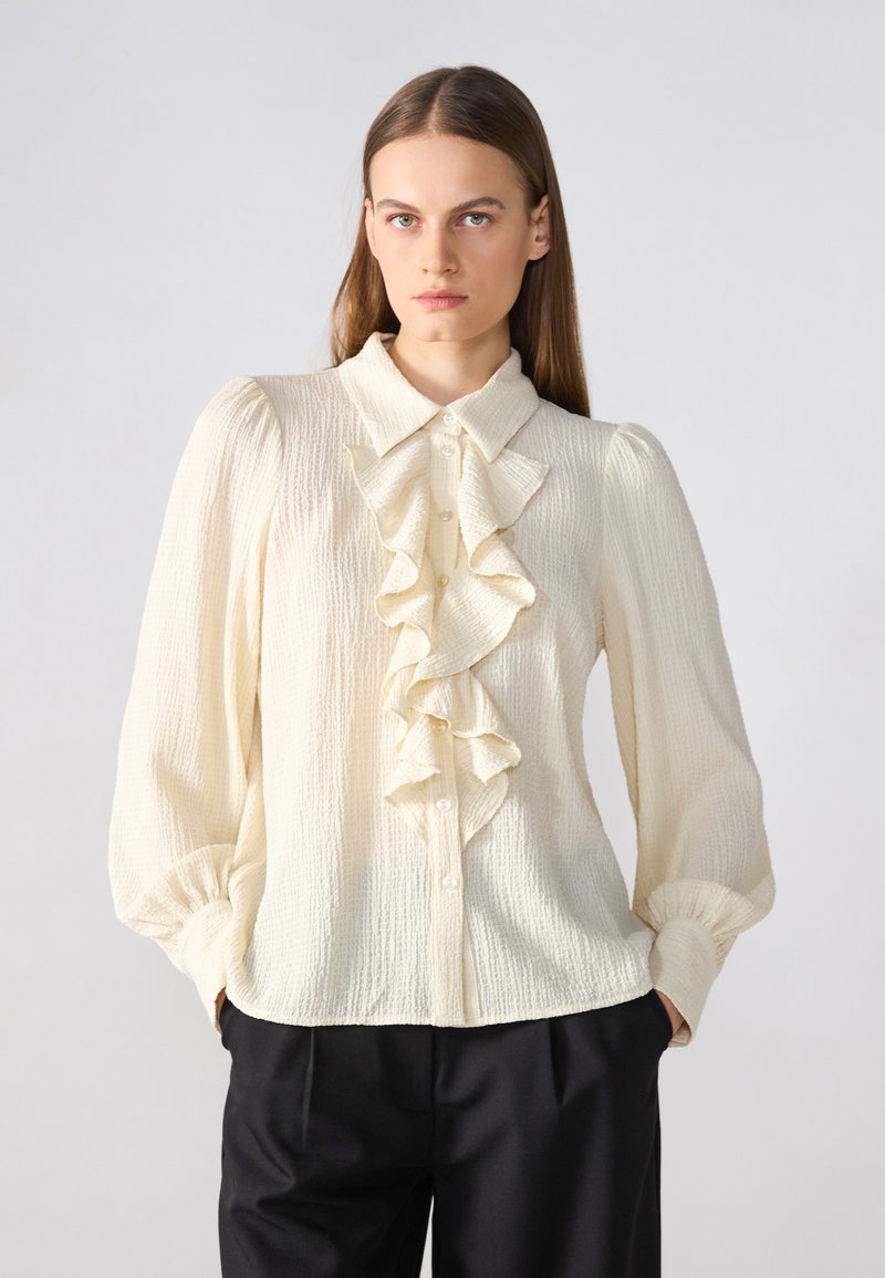 Blusa de color crema, texturizada, con fruncidos en el frente, cuello y mangas largas. Botones en la parte delantera, combinada con pantalones negros.